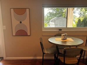 A dining area with a round table, four chairs, a wall-mounted abstract painting, and a window with a view of a shed and trees. - Cozy 1-bedroom apartment in lovely Weaverville with AC (Weaverville)