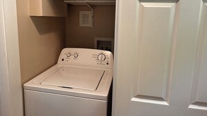 A compact laundry room with a front-loading washing machine, a shelf with cleaning supplies, and a door leading to another room. - Cozy 1-bedroom apartment in lovely Weaverville with AC (Weaverville)