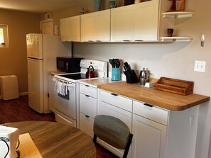 A compact kitchen with white cabinetry, a microwave, an oven, and a kettle on the countertop. There is a chair and a dining table with a wooden top. - Cozy 1-bedroom apartment in lovely Weaverville with AC (Weaverville)