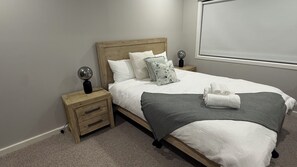 A neatly made bed with a grey throw, white pillows, and a wooden headboard. There are two bedside tables with lamps and a small drawer. - Peninsula Beach house (Mount Eliza)