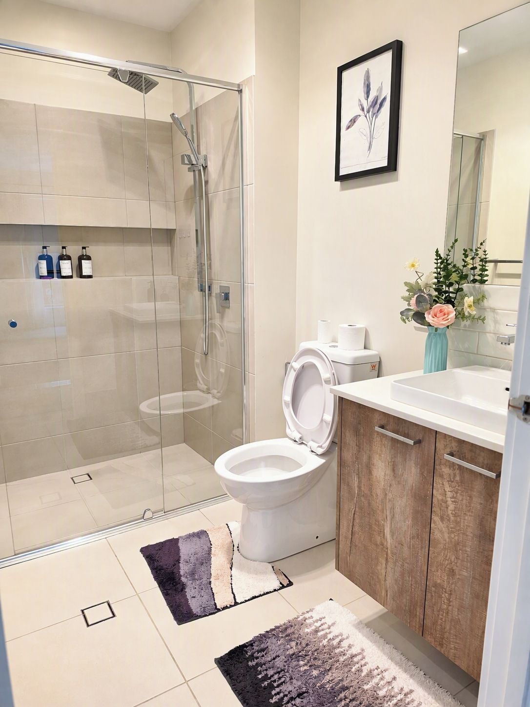 A modern bathroom with a glass shower enclosure, a toilet, a sink with a wooden cabinet, and a framed floral artwork on the wall.