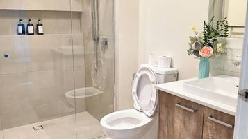 A modern bathroom with a glass shower enclosure, a toilet, a sink with a wooden cabinet, and a framed floral artwork on the wall.