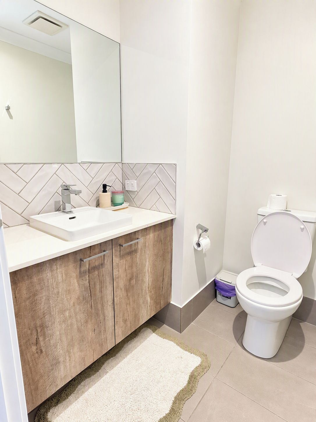 Ein Badezimmer mit weißem Waschbecken, Holzschublade, einem Spiegel, einer Toilette und einer Wandverkleidung mit Fischgrätmuster.