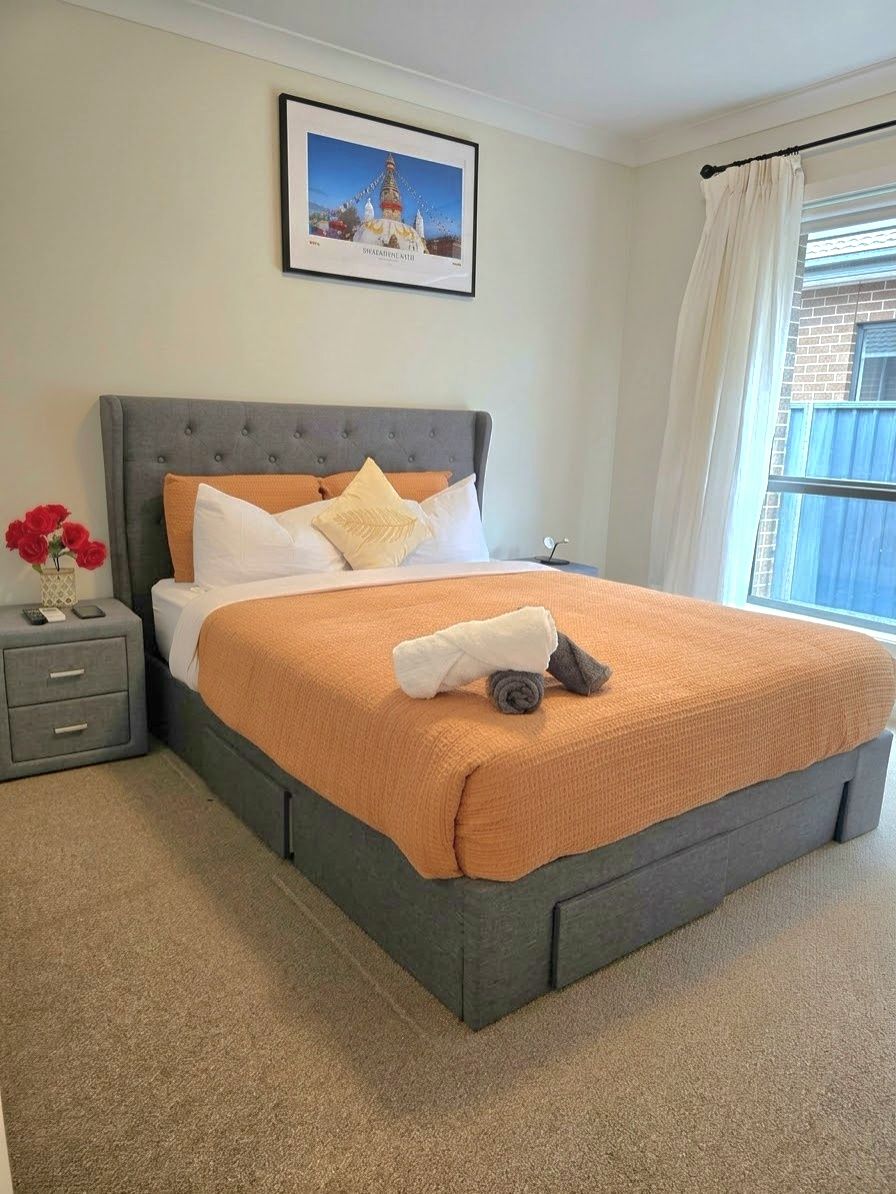 A neatly made bed with a grey headboard, a framed picture above it, and a window with curtains to the right.