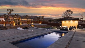 A pool area with a view of the mountains, outdoor seating, and a building with illuminated windows. - JT luxe Compound Pool, Spa, Floodlit Pickleball, Sauna, Dome (Joshua Tree)