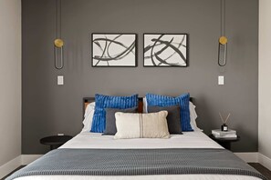 A modern hotel room with a large bed, two wall-mounted artworks, a bedside table with a book and a candle, and a nightstand with a lamp. - JT luxe Compound Pool, Spa, Floodlit Pickleball, Sauna, Dome (Joshua Tree)