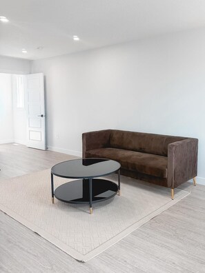 Ein Zimmer mit einem braunen Sofa, einem runden schwarzen Tisch und einem gemusterten Teppich auf einem hellen Holzboden.