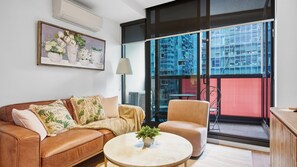 A living room with a brown leather sofa, a round coffee table with a plant, and a large window with a view of buildings. - The Rose Above the City Melbourne CBD (Melbourne)