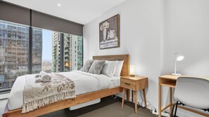 Une chambre moderne avec un grand lit, des tables de chevet, un bureau et une vue sur le paysage urbain grâce à de grandes fenêtres du sol au plafond.