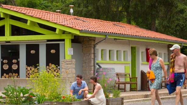 Ein Gebäude mit grüner Fassade und rotem Ziegeldach, ein Gartenbereich mit Pflanzen und Blumen sowie eine hölzerne Bank.