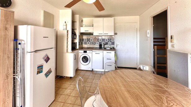 A compact kitchen with a ceiling fan, a refrigerator, a washing machine, and a dining area with a table and chairs. - Apartment canal view with parking - 4 guests (Sète)
