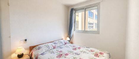 Un dormitorio con cama, mesita de noche, lámpara, ventilador de techo y ventana con cortinas.