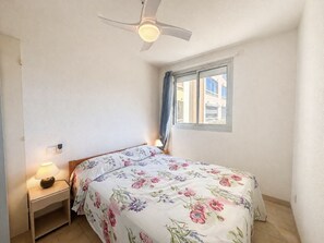 Ein Schlafzimmer mit einem Bett, einem Nachttisch, einer Lampe, einem Deckenventilator und einem Fenster mit Vorhängen.