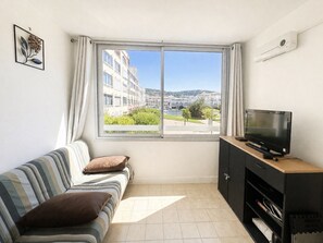 A room with a bed, a TV on a stand, a window with a view of buildings and greenery, and a wall-mounted air conditioner. - Apartment canal view with parking - 4 guests (Sète)