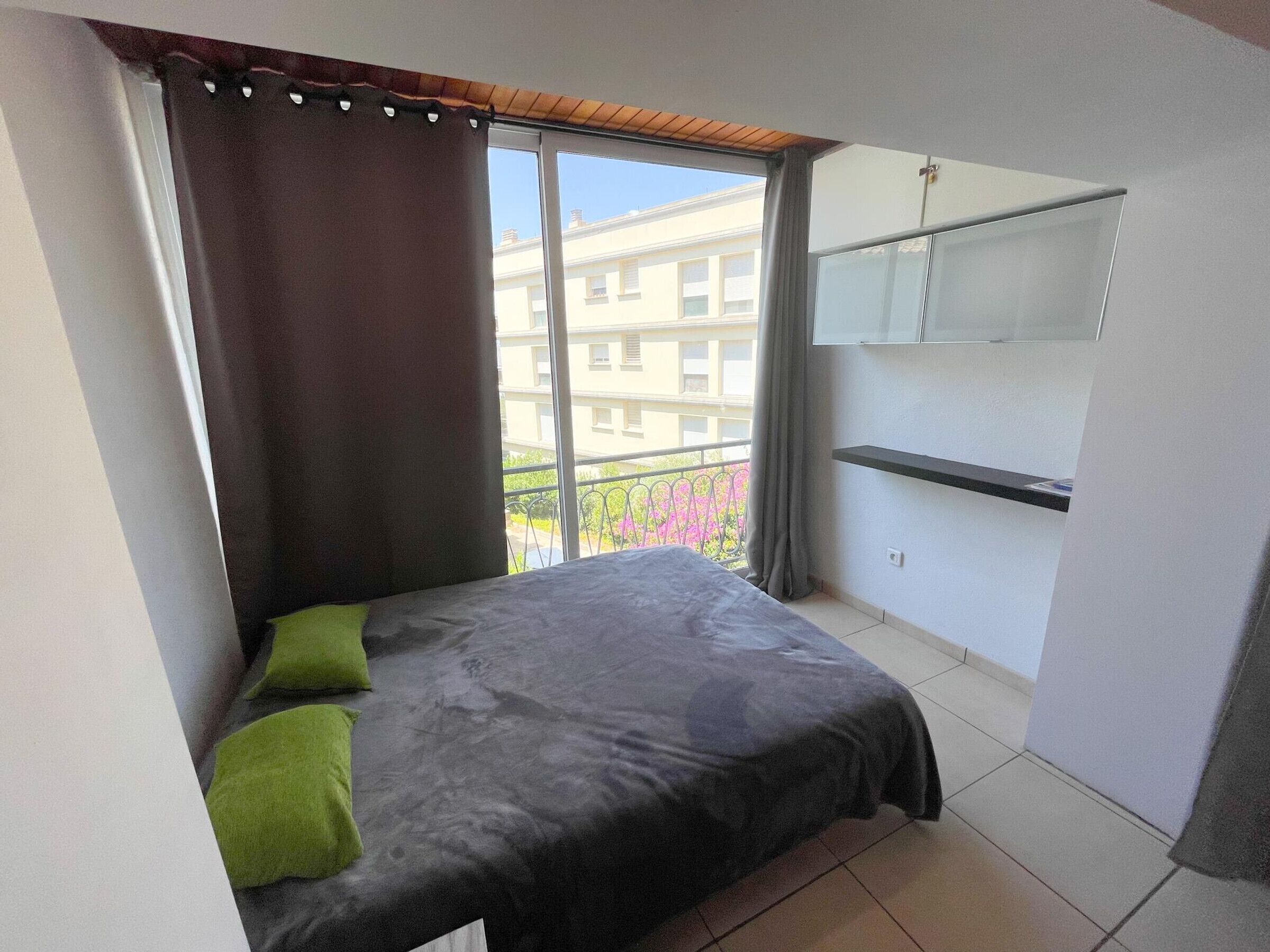 A bedroom with a bed, a window with curtains, a balcony, and a small shelf.