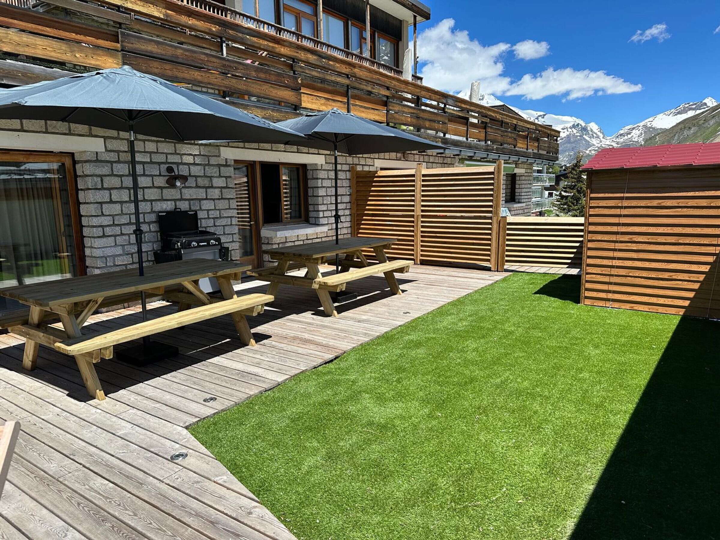 Ein Patio mit Holztischen und Sonnenschirmen, ein Steinhaus mit Grill und Blick auf schneebedeckte Berge.