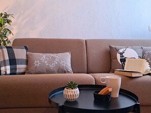 Ein beiges Sofa mit gemusterten Kissen, ein kleiner Topfpflanze, ein Tablett mit Tasse und Keksen sowie ein Buch auf einem Beistelltisch.