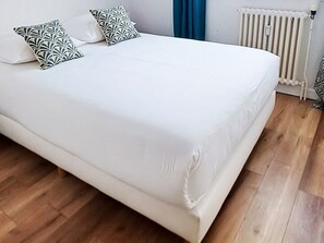 Ein Bett mit weißer Bettwäsche und gemusterten Kissen, ein Heizkörper und Holzdielen.