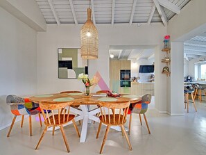 A dining area with a round wooden table, six chairs, a hanging light fixture, and a kitchen in the background.