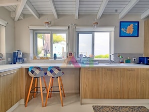 A modern kitchen with wooden cabinets, two bar stools, a coffee machine, and a view of the outdoors. - House with panoramic sea view, pool - 6 guests (Saint-Martin)