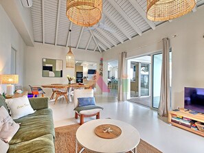 A spacious living room with a large sofa, a coffee table, and a television. There is a dining area with a table and chairs, and a kitchen in the background. - House with panoramic sea view, pool - 6 guests (Saint-Martin)