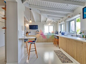 A modern kitchen with wooden cabinets, a central island, bar stools, and a large window providing natural light. - House with panoramic sea view, pool - 6 guests (Saint-Martin)