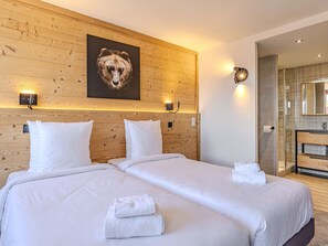 Et hotelværelse med to senge, en træsenggavl, et billede af en bjørn, væglamper, et badeværelse med en glasbrusekabine og et spejl.