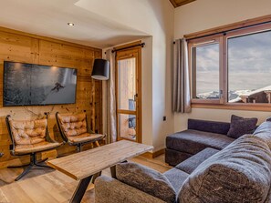 Ein gemütliches Zimmer mit einer Holzwand, einem Wand-Fernseher, Ledersesseln, einer Couch und einem großen Fenster mit Blick auf Schnee.