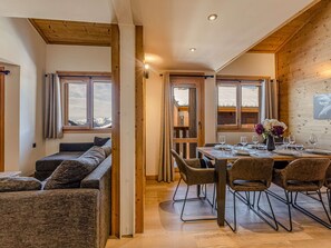 A dining area with a wooden table set for four, flanked by two windows with curtains, and a sofa in the adjacent room. - Charming duplex in the heart of Belle Plagne - 8 guests (Belle Plagne)