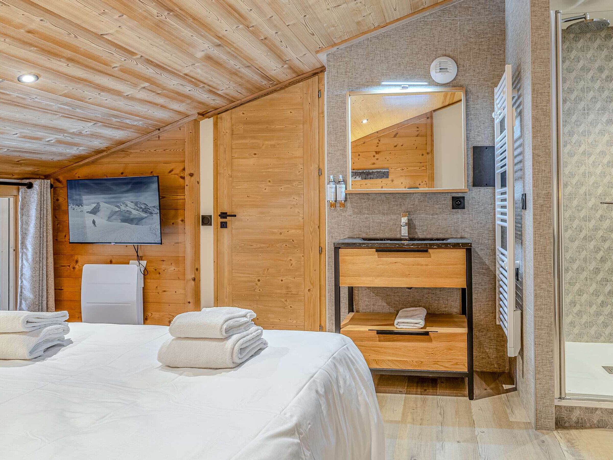 Une chambre d’hôtel avec un plafond en bois, un lit avec du linge de maison blanc, un téléviseur fixé au mur, une coiffeuse en bois avec un miroir et une salle de bain avec douche.