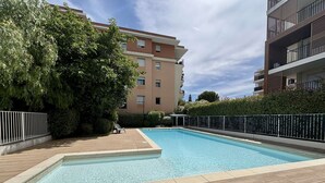 En swimmingpool med fliseterrasse, omgivet af et hegn og træer, med en høj bygning i baggrunden.