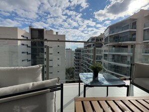 Ein Balkon mit Blick auf benachbarte Gebäude, einem Glastisch und einer Couch.