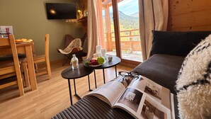 Um quarto aconchegante com um sofá, uma mesa de jantar com cadeiras, uma pequena mesa lateral com um livro e copos, e uma televisão na parede.