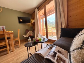 Ein gemütliches Zimmer mit einer Couch, einem Esstisch mit Stühlen, einem kleinen Beistelltisch mit Buch und Gläsern sowie einem an der Wand befestigten Fernseher.