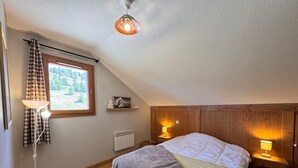 Quarto com teto inclinado, uma cama com cobertor, duas luminárias de cabeceira, um radiador e uma janela com cortinas.