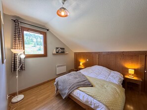 Ein Schlafzimmer mit schrägen Wänden, einem Bett mit Decke, zwei Nachttischlampen, einem Heizkörper und einem Fenster mit Vorhängen.