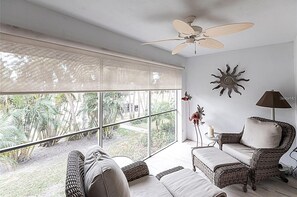 Ein Zimmer mit Deckenventilator, großen Fenstern, Korbmöbeln und einem Sonnendekor an der Wand.