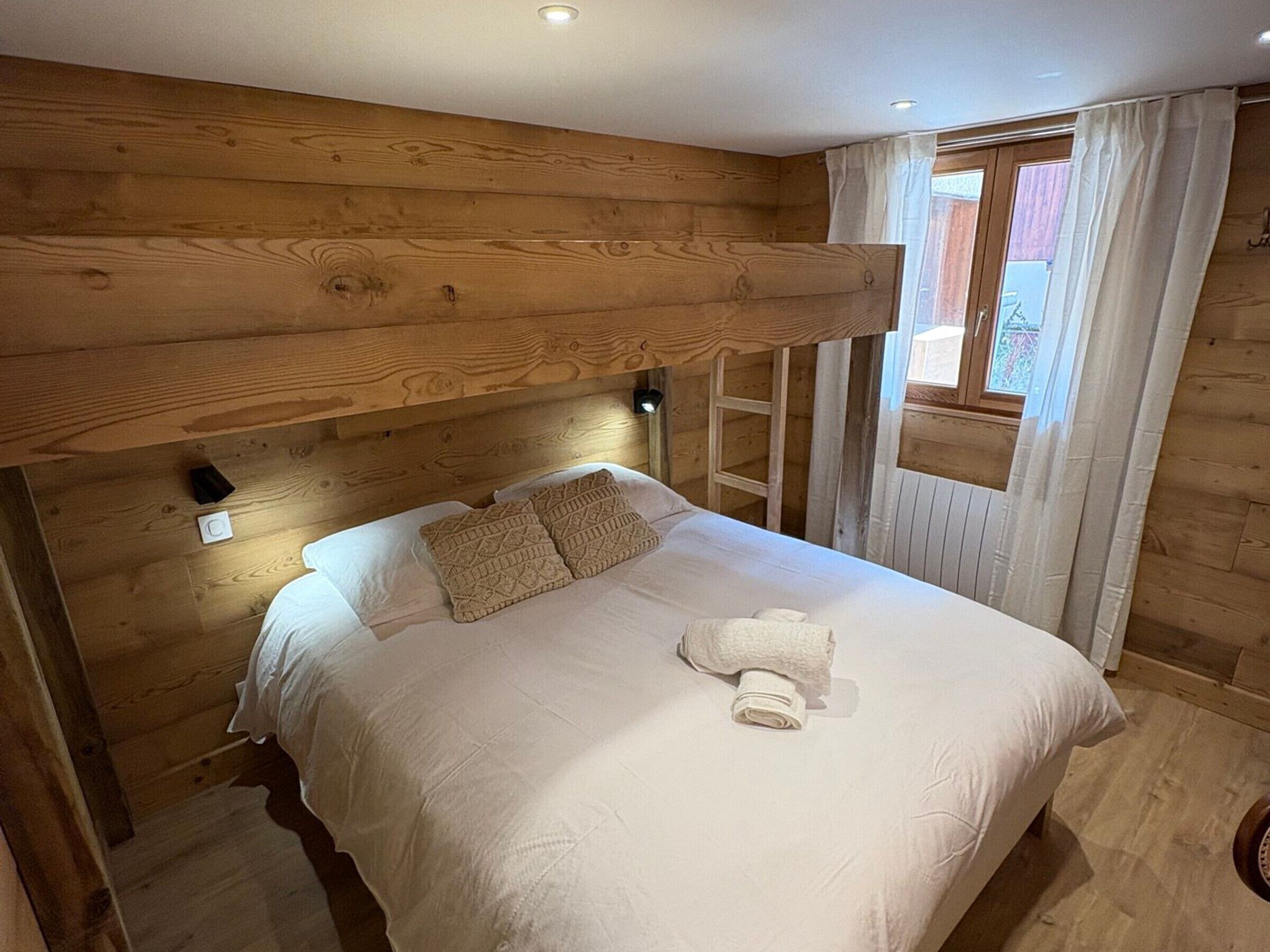 Una habitación tipo cabaña de madera con una cama nido, una escalera y una ventana con cortinas.