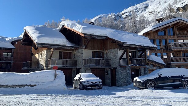 Una cabaña de madera con paredes de piedra, un balcón y un coche cubierto de nieve.