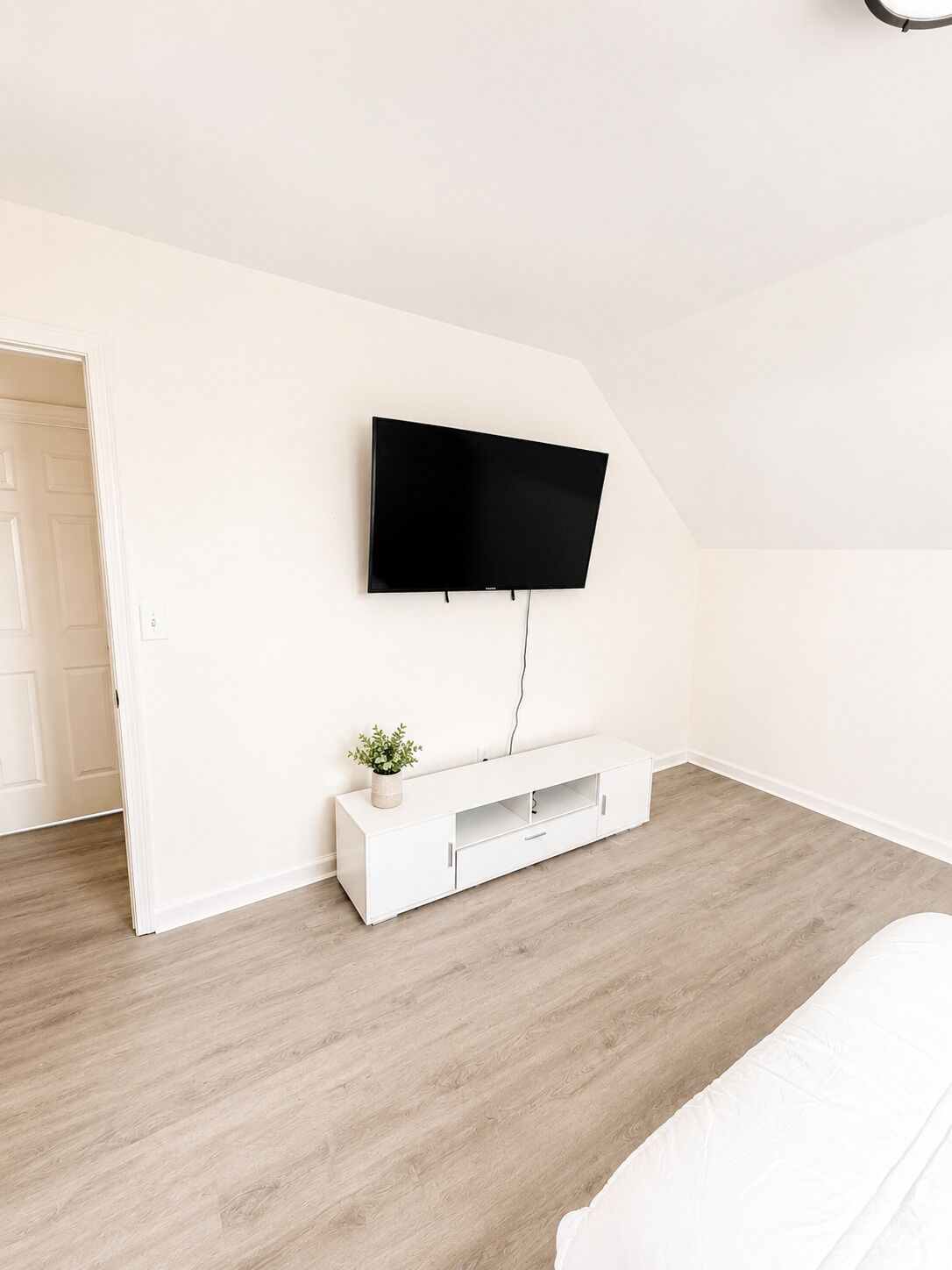 Una habitación con televisión de pantalla plana montada en la pared, una mesita consola blanca con una planta y suelo de madera.