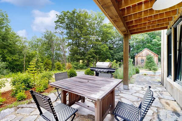 An outdoor patio with a wooden table and chairs, a grill, and a stone pathway leading to a house.
