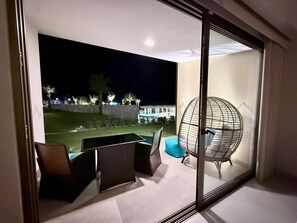 En balkon med glasdør, siddepladser med stole og udsigt til en bygning og palmer om natten.