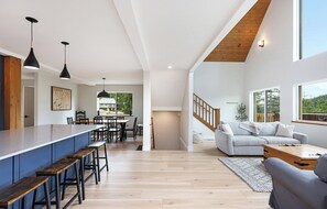 A modern kitchen with a long counter, dining area with chairs, a staircase, and a living room with a sofa and a view of the outdoors. - Ski Bunny Betsy's Chalet | Family  and  Pet-Friendly Mountain Retreat  | Hot Tub | Mountain Views | 5 Minutes to the Lifts (Mount Washington)
