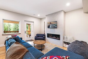 A modern living room with a sectional sofa, a fireplace, and a large window overlooking a deck. - Ski Bunny Betsy's Chalet | Family  and  Pet-Friendly Mountain Retreat  | Hot Tub | Mountain Views | 5 Minutes to the Lifts (Mount Washington)