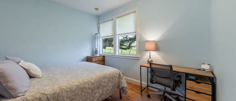 A bedroom with a bed, a desk with a chair, a window with blinds, and a lamp.