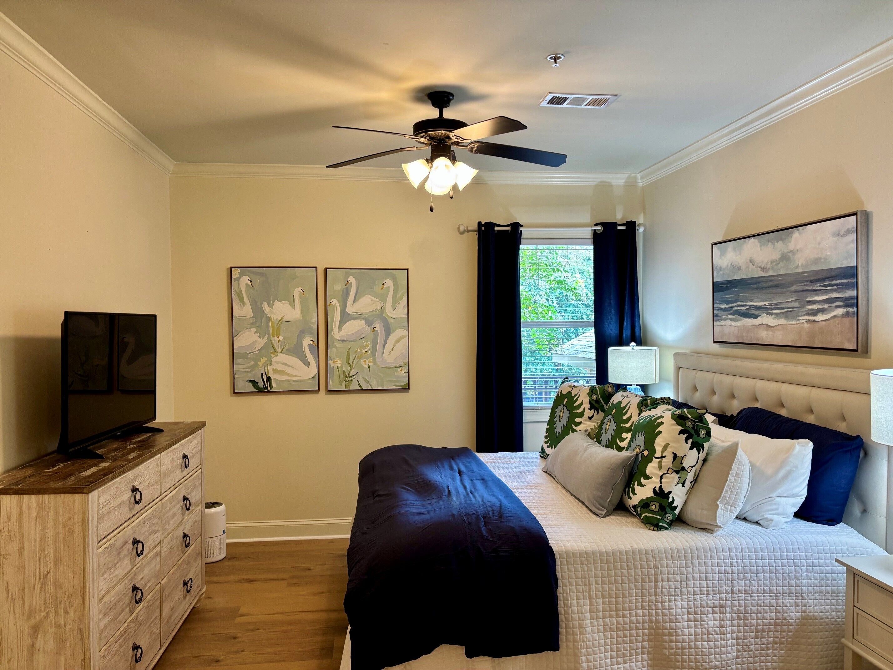 A bedroom with a bed, a ceiling fan, artwork, a television, and a window with curtains.