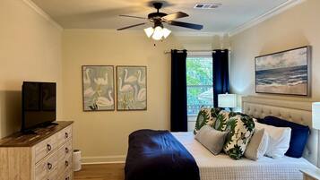 A bedroom with a bed, a ceiling fan, artwork, a television, and a window with curtains.