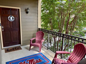 A porch with a dark wooden door, a red chair, a blue mat with 'Miss' written on it, and a metal railing.