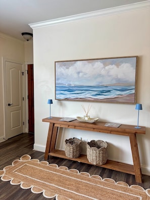 A hallway with a wooden console table, a painting of a seascape, two lamps, and woven baskets.
