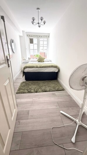 A narrow hallway with a bed, a fan, and a window with a view of buildings. - Apartment Sleeps 11 with Air Conditioning (London)